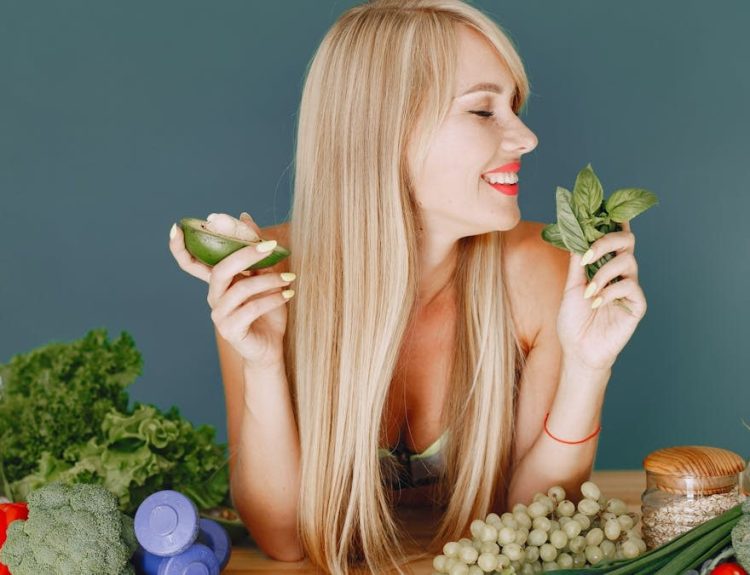 A smiling woman holding avocado and basil, surrounded by fresh vegetables and fruits for healthy eating.